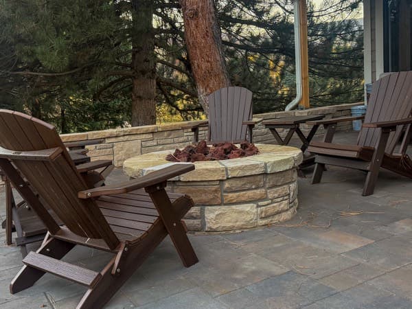 Round stone fire pit with Adirondack chairs on a patio among pine trees