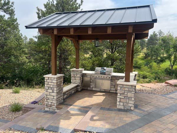 Timber pavilion with a metal roof over a stone outdoor kitchen and paver patio